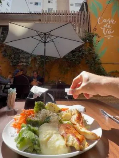 Mão temperando prato com mural Casa de Vó ao fundo